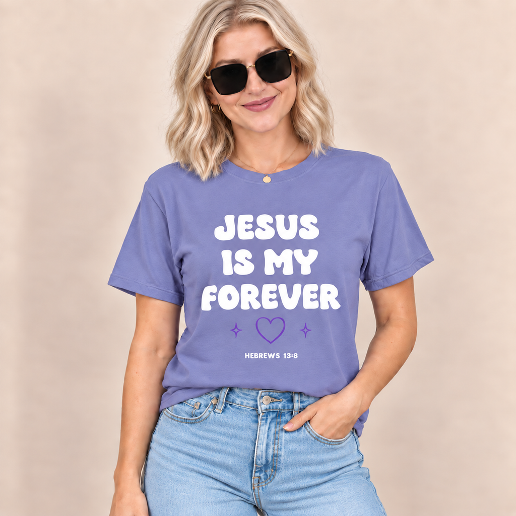 A woman wearing a lavender-purple graphic t-shirt with the text 'JESUS IS MY FOREVER' in white and a small purple heart, paired with light-wash blue jeans against a neutral beige background.
