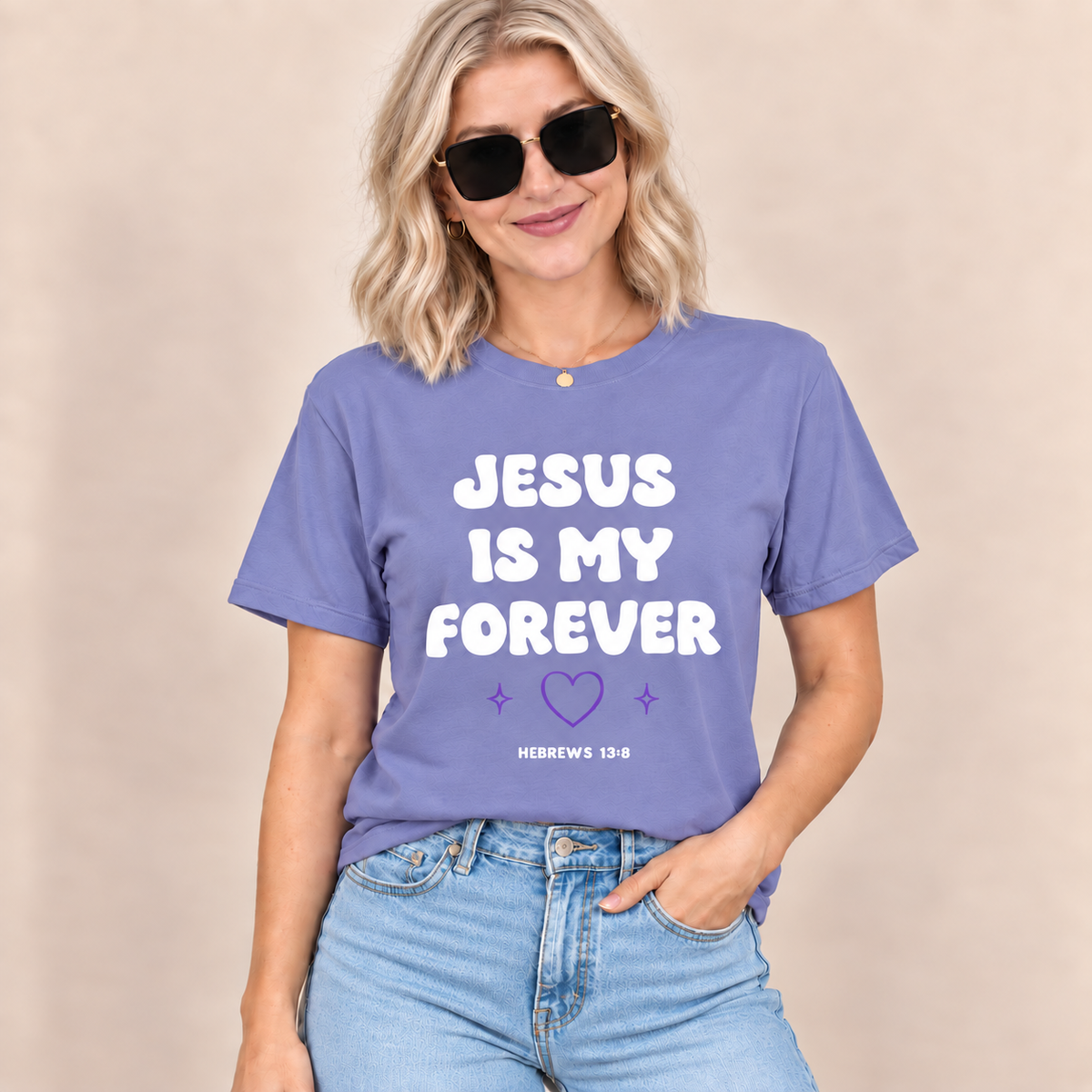 A woman wearing a lavender-purple graphic t-shirt with the text 'JESUS IS MY FOREVER' in white and a small purple heart, paired with light-wash blue jeans against a neutral beige background.